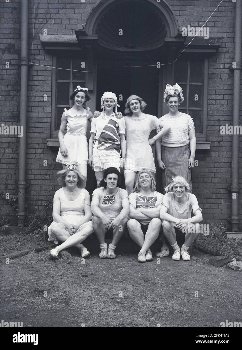 Anni '50, storico, in piedi fuori ad un'entrata ad un edificio, un gruppo di uomini locali - 'The Heavy Gang', Inghilterra, Regno Unito. Vestiti da donna e stanchi di Union Jacks, eseguiranno il loro "Gang Show" nelle celebrazioni del giorno di maggio. Il giorno di maggio era un'antica tradizione che celebrava l'arrivo della primavera e dell'estate e gli artisti si vestirebbero e rievocavano il folklore antico e le storie danzando intorno ad un palo di legno alto, un maypole, un'attività popolare. Foto Stock