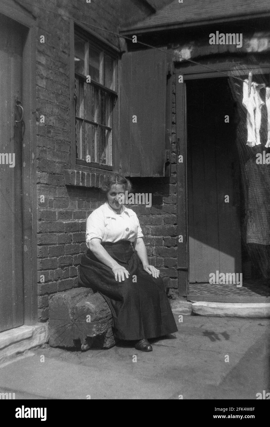 In una giornata di sole brillante, una signora che indossa una lunga gonna di stoffa pesante, tipica dell'epoca, seduta su una piccola panchina di tronchi, nel cortile chiuso di una proprietà vittoriana, Cleethorpes, Inghilterra, Regno Unito, intorno agli anni '30. Foto Stock