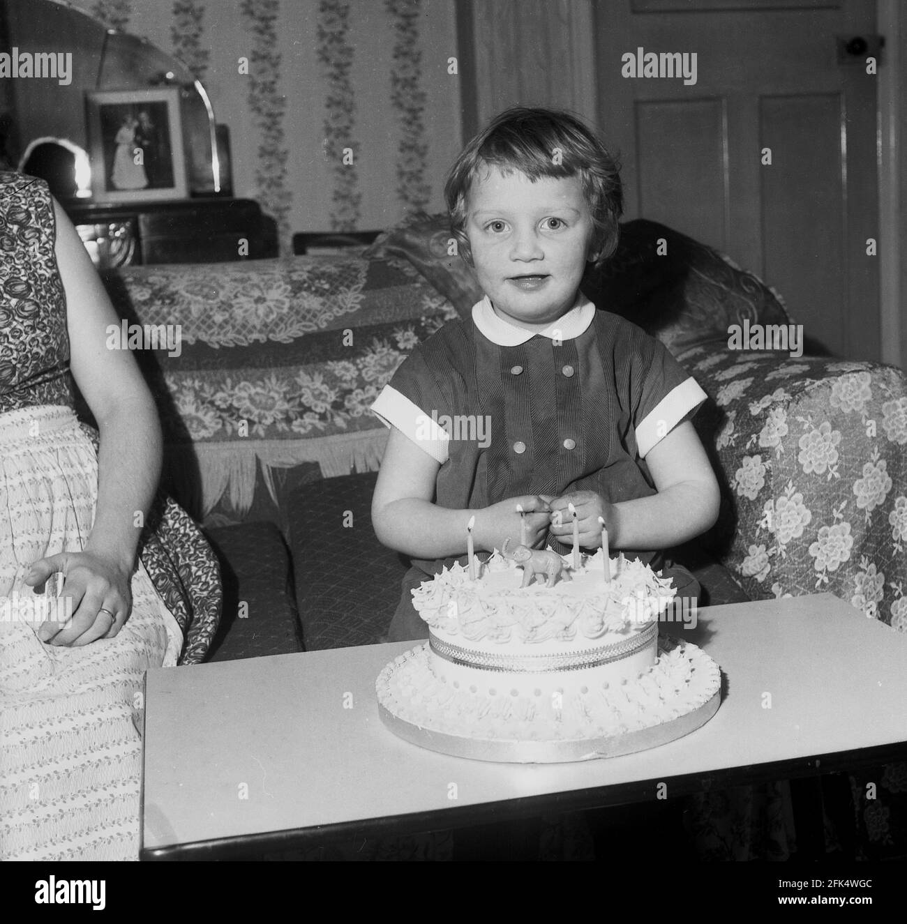 anni '60, storico, su un divano in una stanza anteriore, una bambina seduta  con sulla sua torta di compleanno, con quattro candele accese e un piccolo  elefante sulla parte superiore. Sembra più