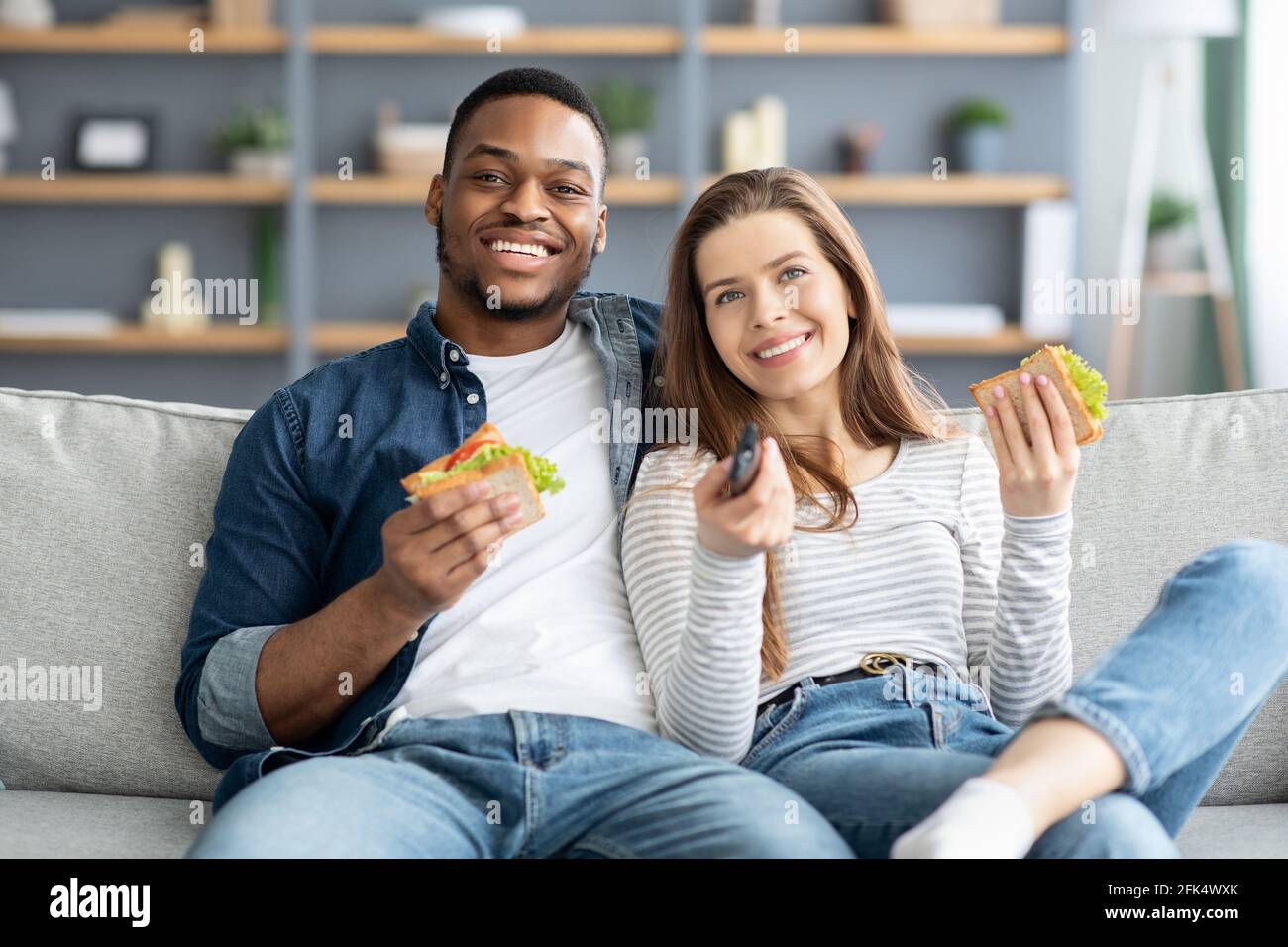 Happy giovane coppia multiculturale guardare la TV e mangiare panini a. Casa Foto Stock