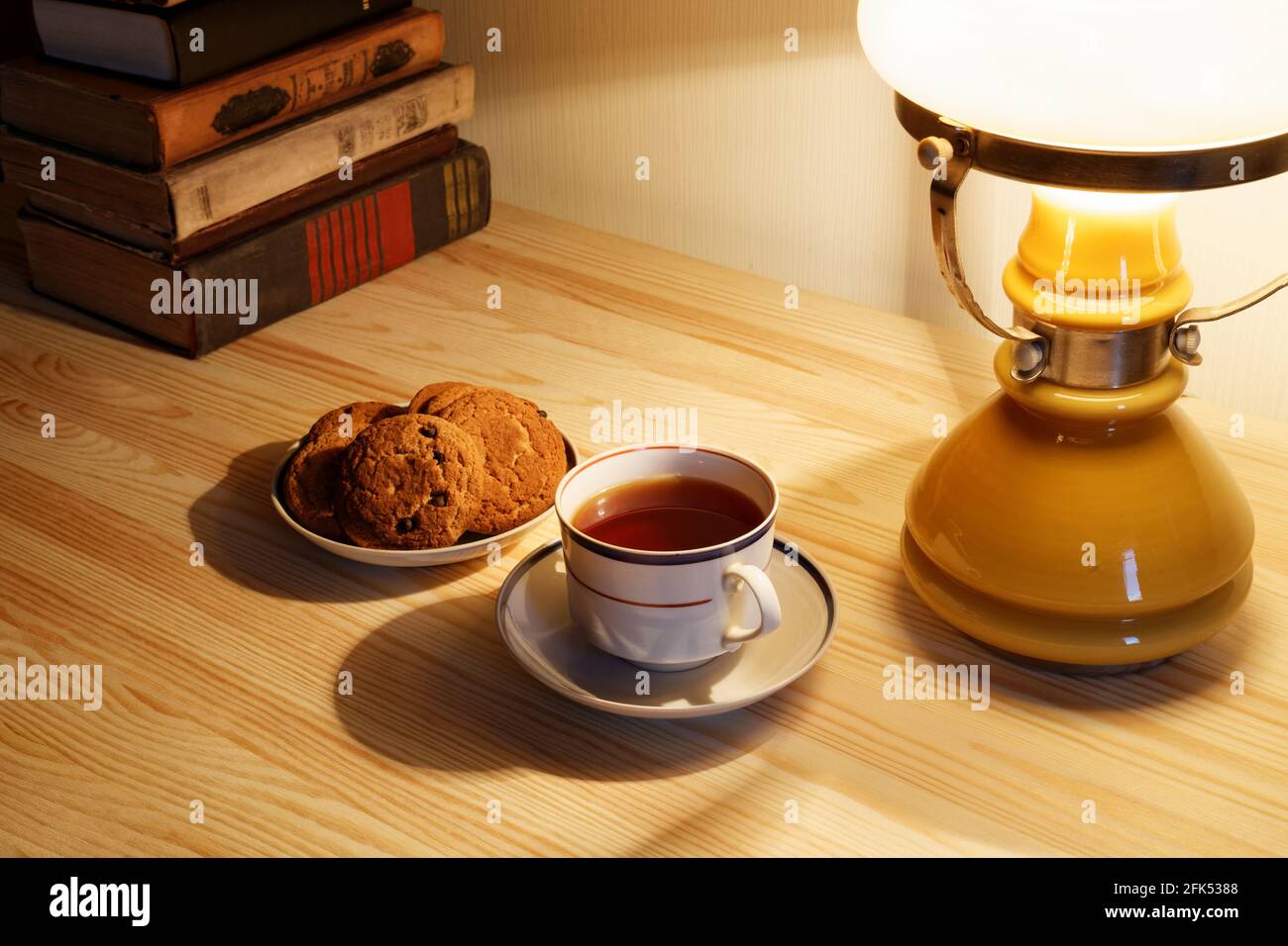 Tazza di tè e pila di vecchi libri sul tavolo con lampada a luce calda Foto Stock