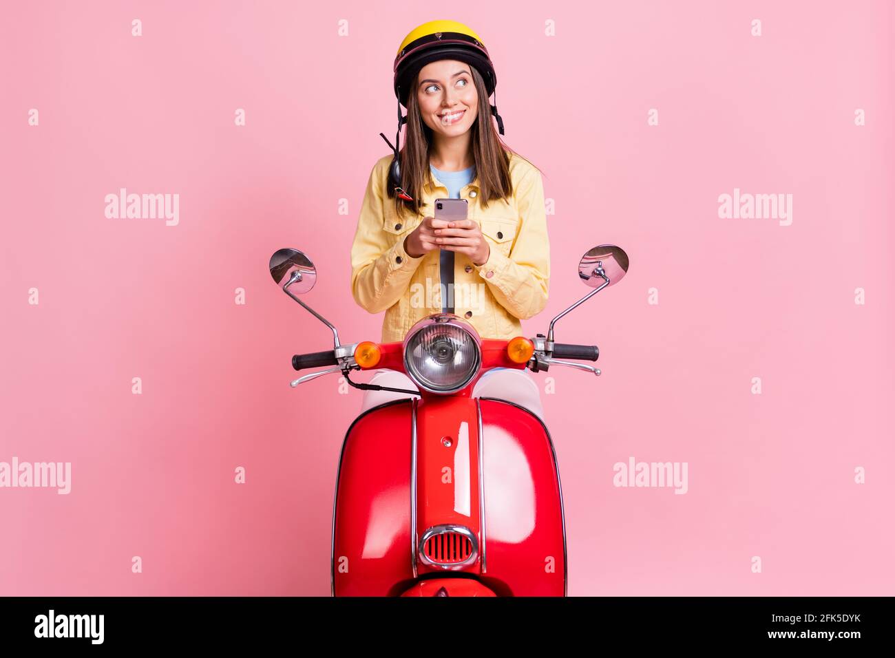 Foto di giovane curiosa ragazza felice guardare copyspace tenere telefono indossare il casco giallo ride moto rosso isolato di colore rosa sfondo Foto Stock