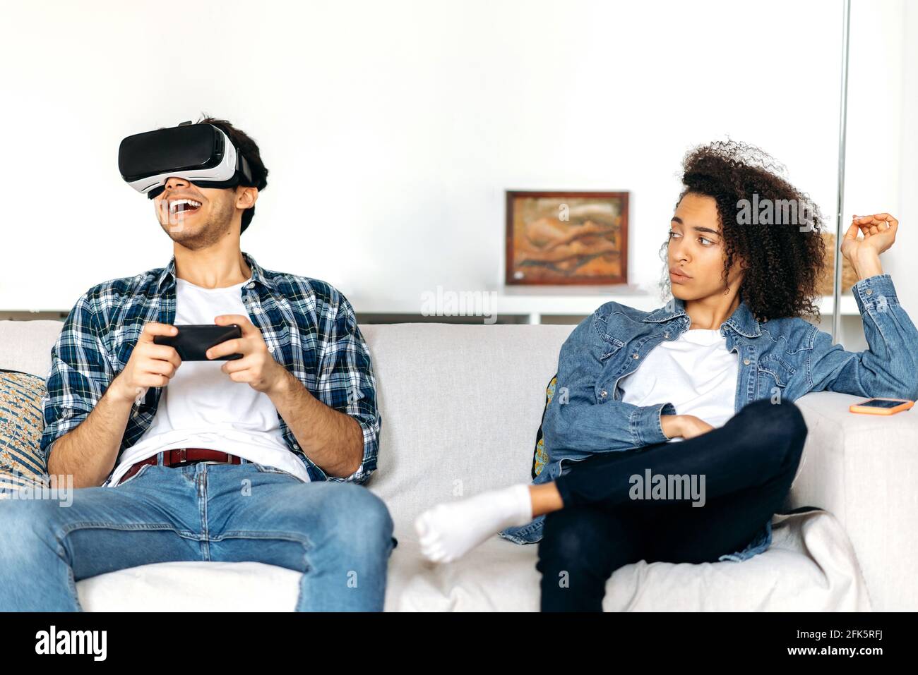 La coppia sposata è seduta sul divano, il ragazzo eccitato utilizza gli occhiali per smartphone e VR, gioca a giochi virtuali, la ragazza afroamericana si annoia e lo guarda indignato Foto Stock