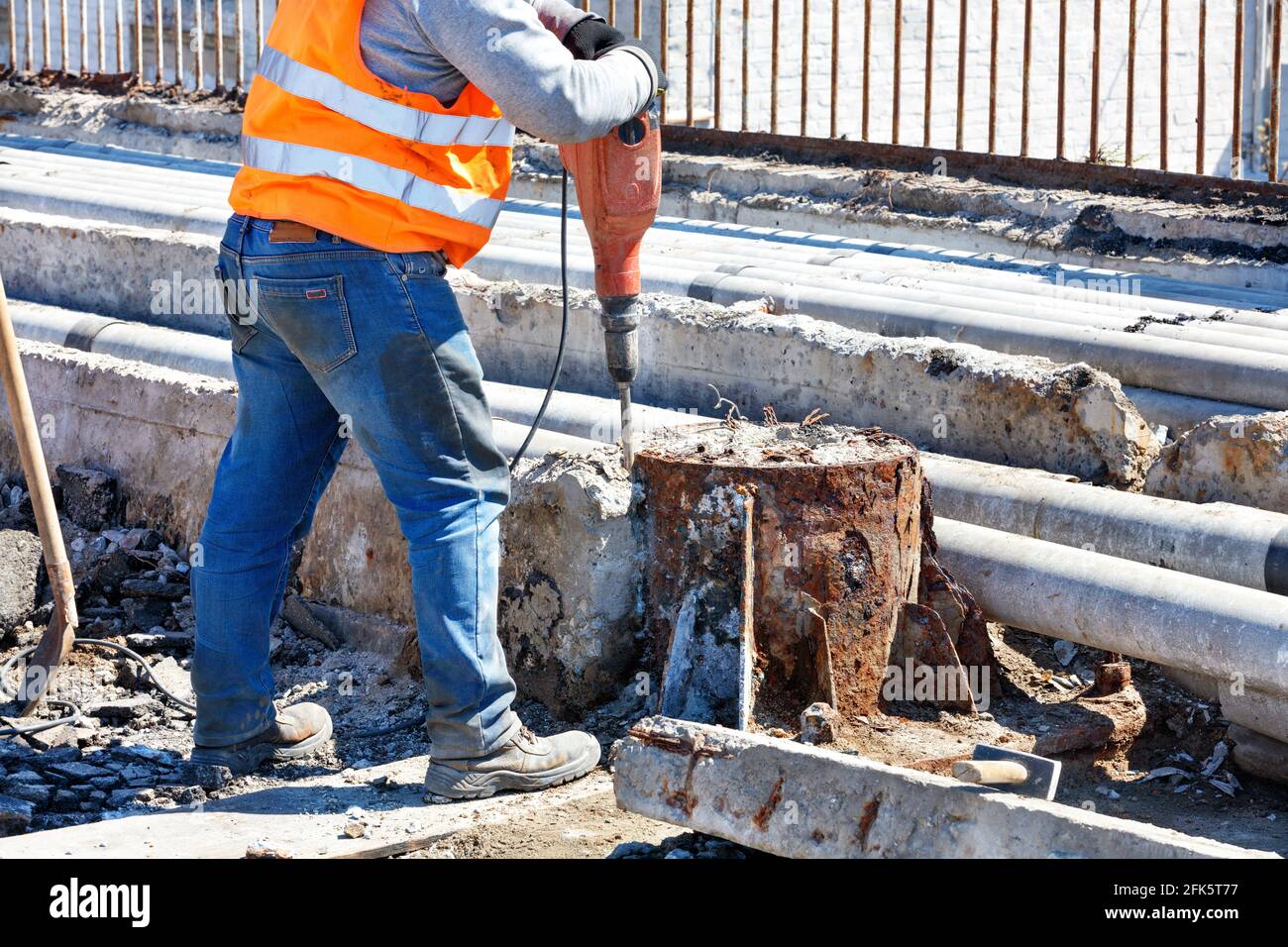 Un lavoratore stradale in un giubbotto riflettente arancione lavora con un martello pneumatico per distruggere una vecchia struttura in cemento armato intorno a un supporto arrugginito. Spazio di copia. Foto Stock