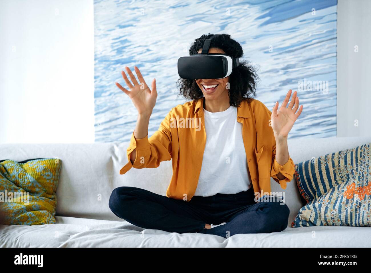 Gioco nel mondo virtuale. Entusiasta ragazza afro-americana con capelli  ricci siede sul divano in abiti