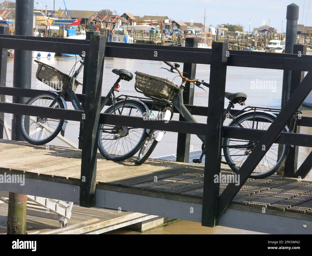 Pedala verso la tua barca: Due biciclette parcheggiate su un molo di legno a Walberswick sul fiume Blyth con il lungomare di Southwold Harbour in lontananza. Foto Stock