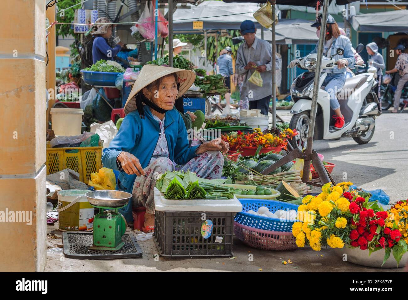 Commerciante vietnamita che indossa cappello conico che vende verdure e fiori sul lato della strada, Hoi An, Vietnam Foto Stock