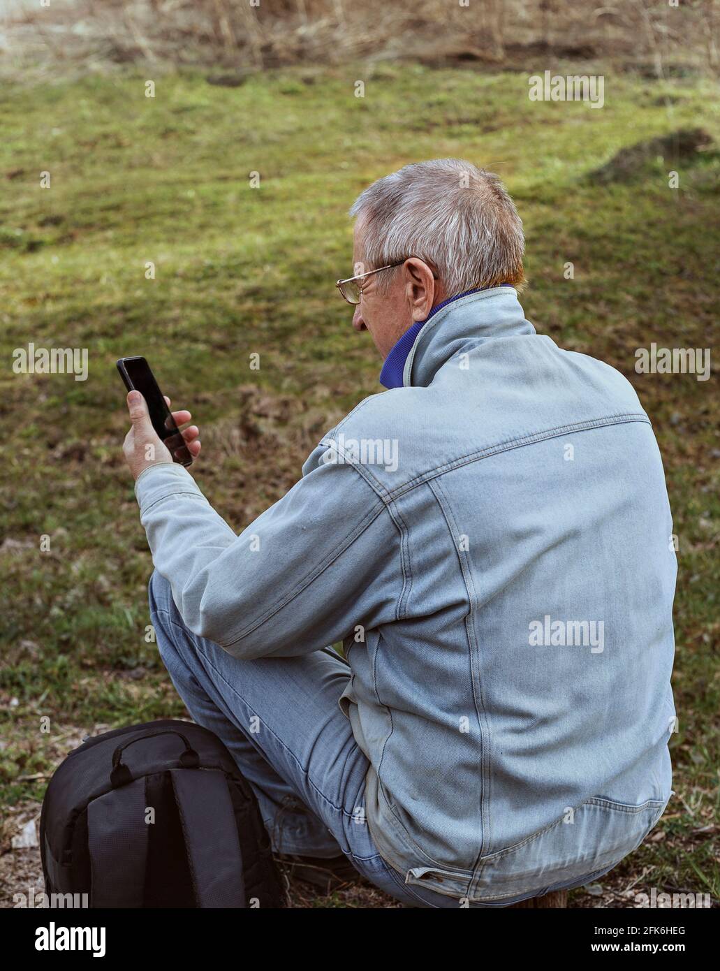 L'uomo anziano in giacca grigia si siede a terra e guarda il suo smartphone. Lo zaino per i viaggi si trova accanto. Foto Stock