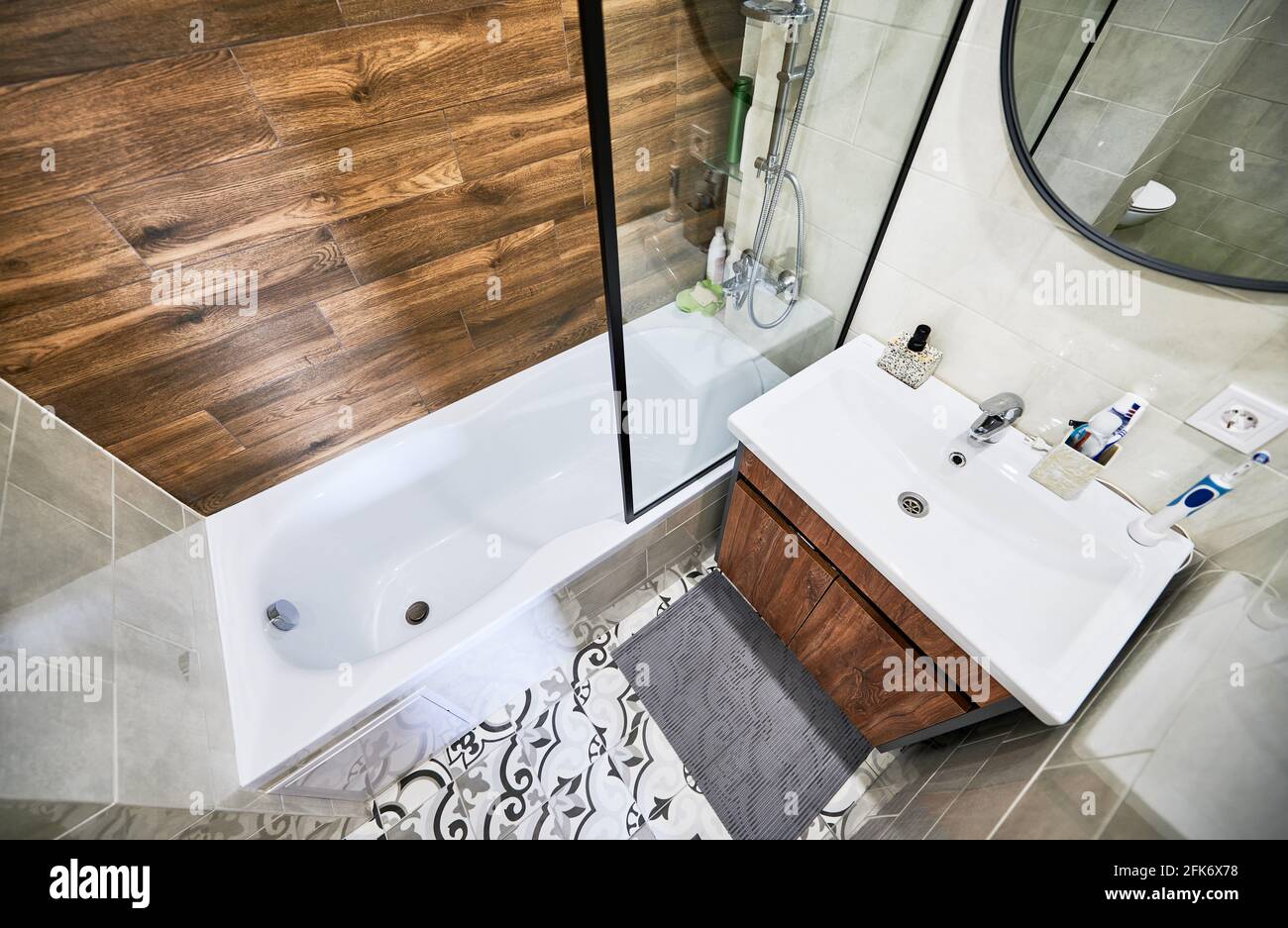 Vista dall'alto della parte bagno con vasca ad angolo retto separata dal lavandino da uno schermo trasparente. Piastrelle da parete che imitazione del legno coincidono con mobili e contrastano con il colore bianco di base. Foto Stock