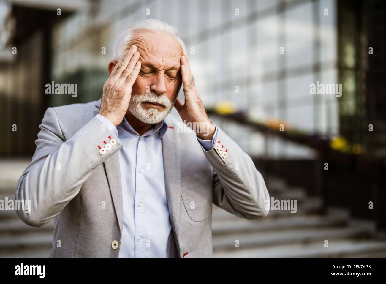 L'uomo d'affari anziano è stanco di lavoro. È in piedi davanti alla costruzione dell'azienda ed ha mal di testa. Foto Stock