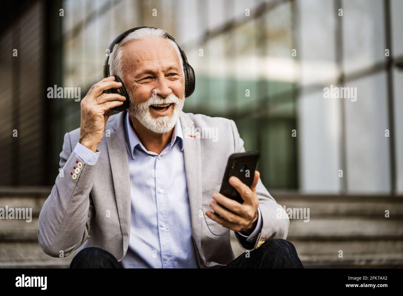Felice uomo d'affari senior è rilassante dopo il lavoro di fronte alla costruzione dell'azienda. Sta ascoltando musica con le cuffie. Foto Stock