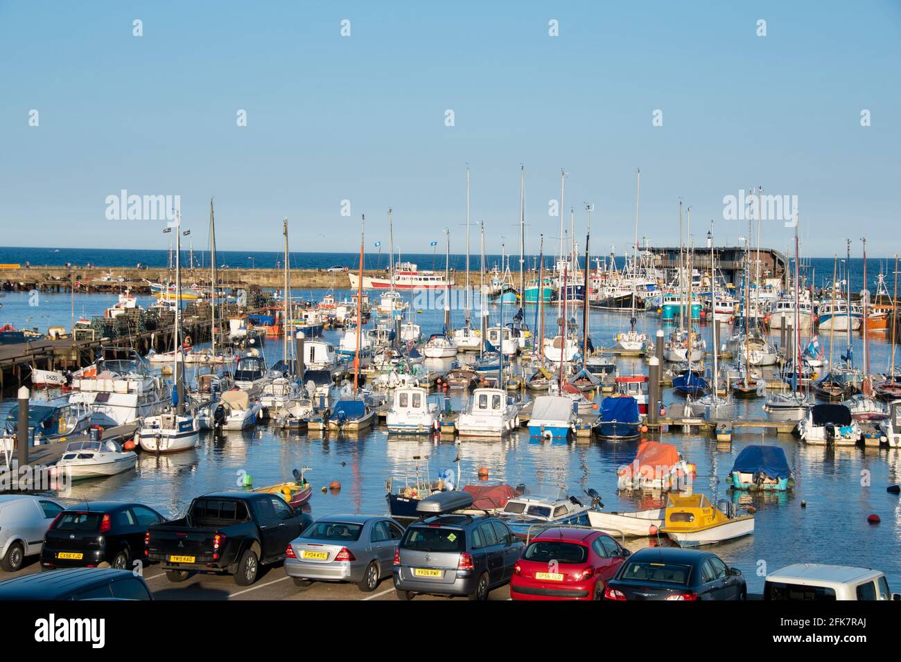 Yorkshire, UK – 10 ago 2017 : una fila di auto si affaccia su un porto trafficato pieno di barche a vela colorate, la flotta di Bridlington Harbour Foto Stock