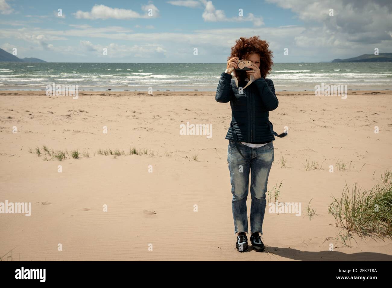 Bella giovane donna con capelli rossi ricci che tiene una macchina fotografica giocattolo di legno su sfondo di spiaggia di sabbia vuota. Concetto di fotografia divertente. Foto Stock