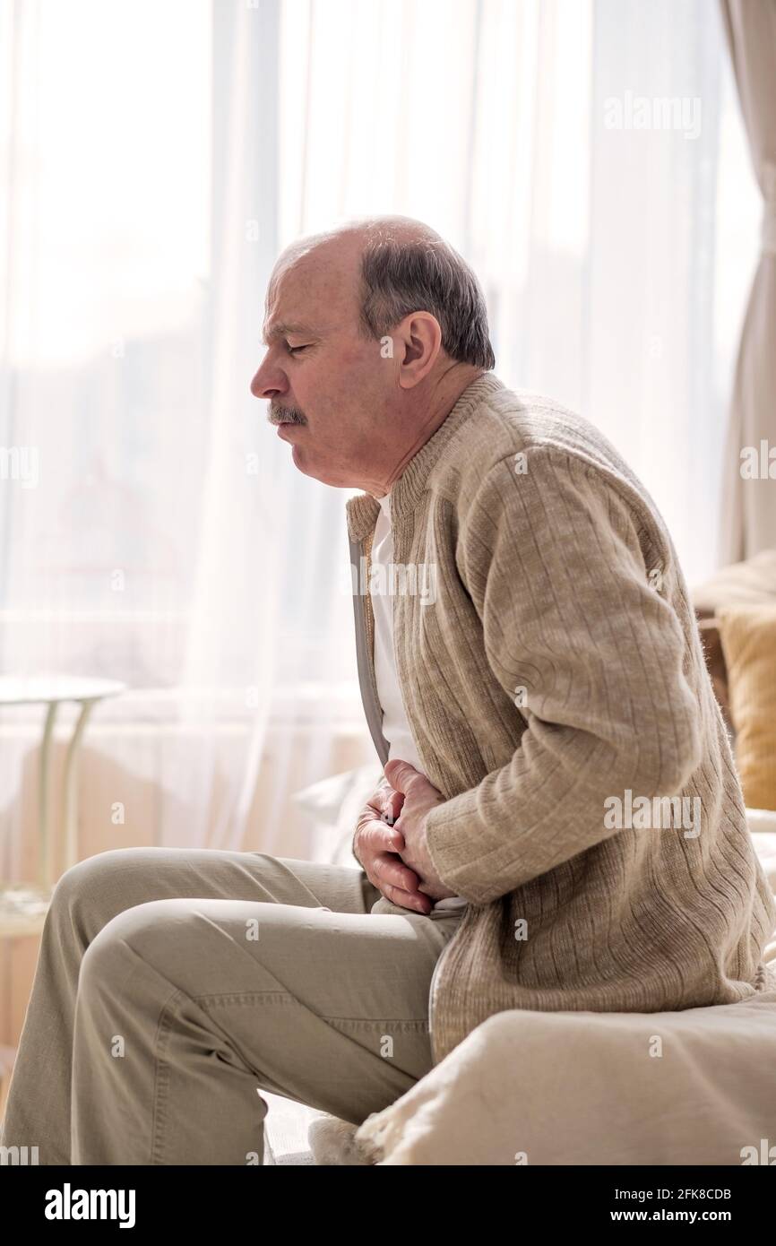 Uomo anziano che ha dolore dello stomaco seduto a casa da solo. Foto Stock