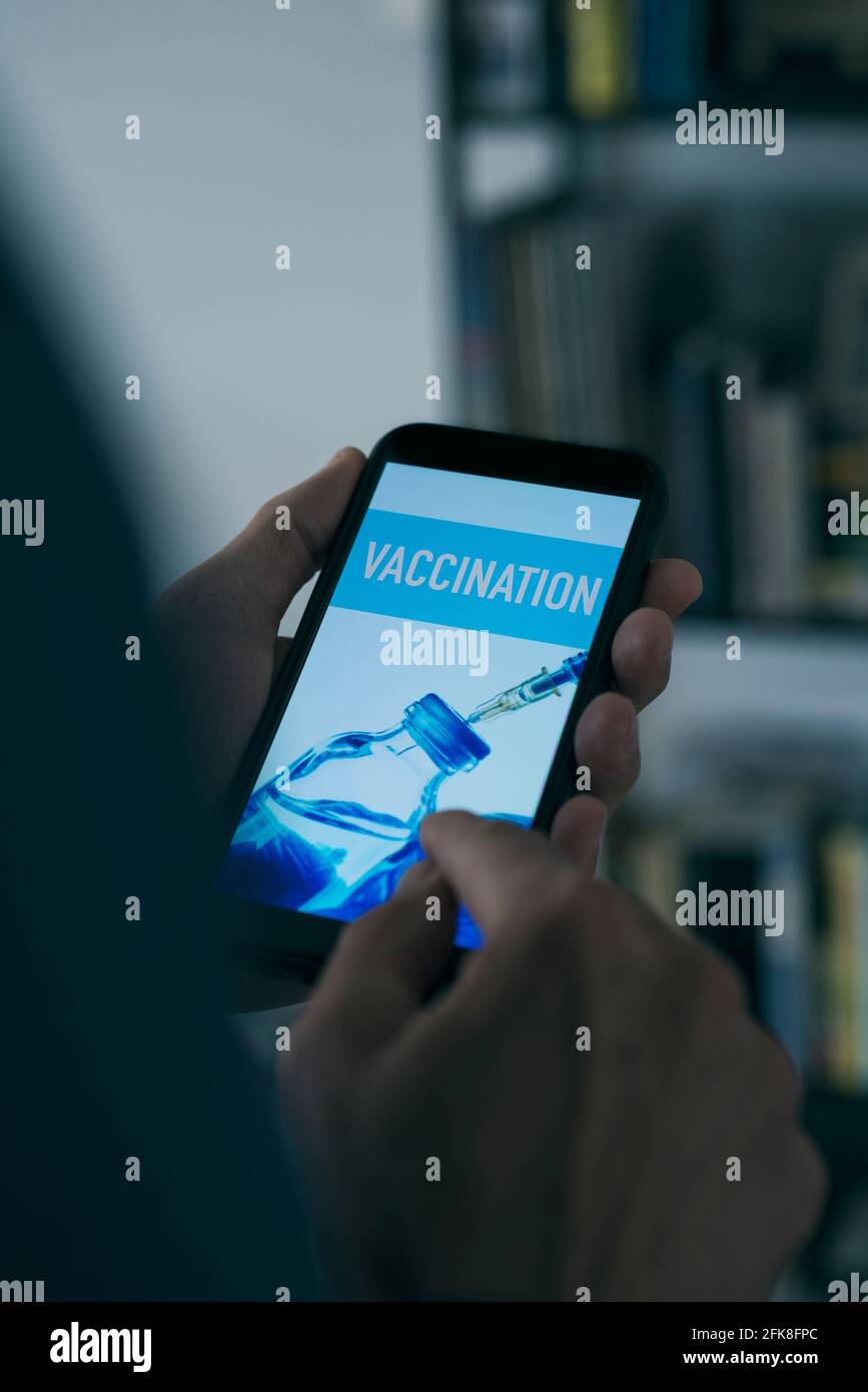 primo piano di un giovane caucasico, nel suo salotto, guardando il suo smartphone, con la vaccinazione di testo nel suo schermo Foto Stock