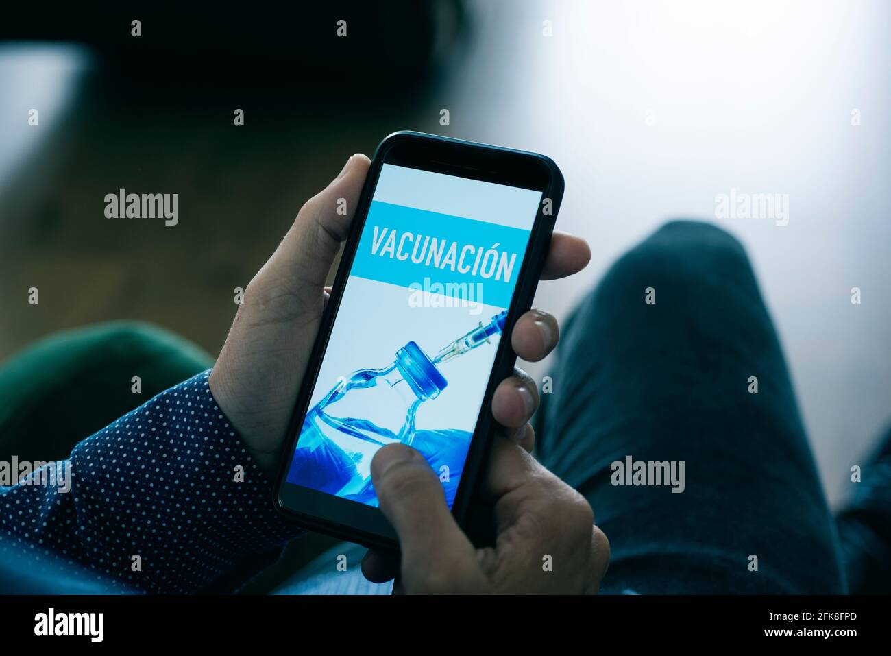 primo piano di un giovane caucasico, seduto in soggiorno, guardando il suo smartphone, che legge il testo vaccinazioni scritto in spagnolo nel suo sc Foto Stock