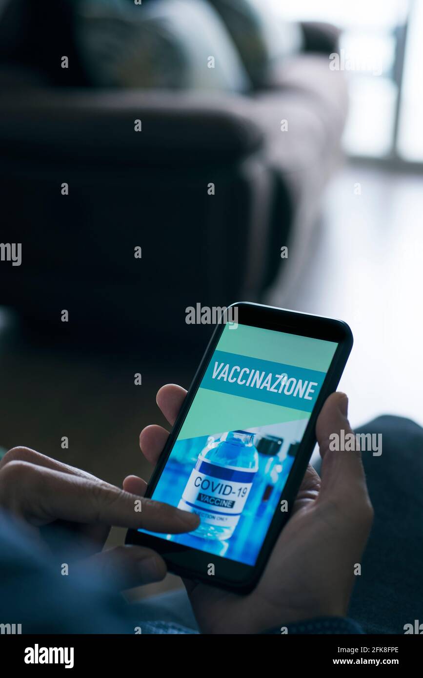 closeup di un giovane caucasico, seduto in soggiorno, guardando il suo smartphone, che legge il testo vaccinazioni scritto in italiano nel suo sc Foto Stock