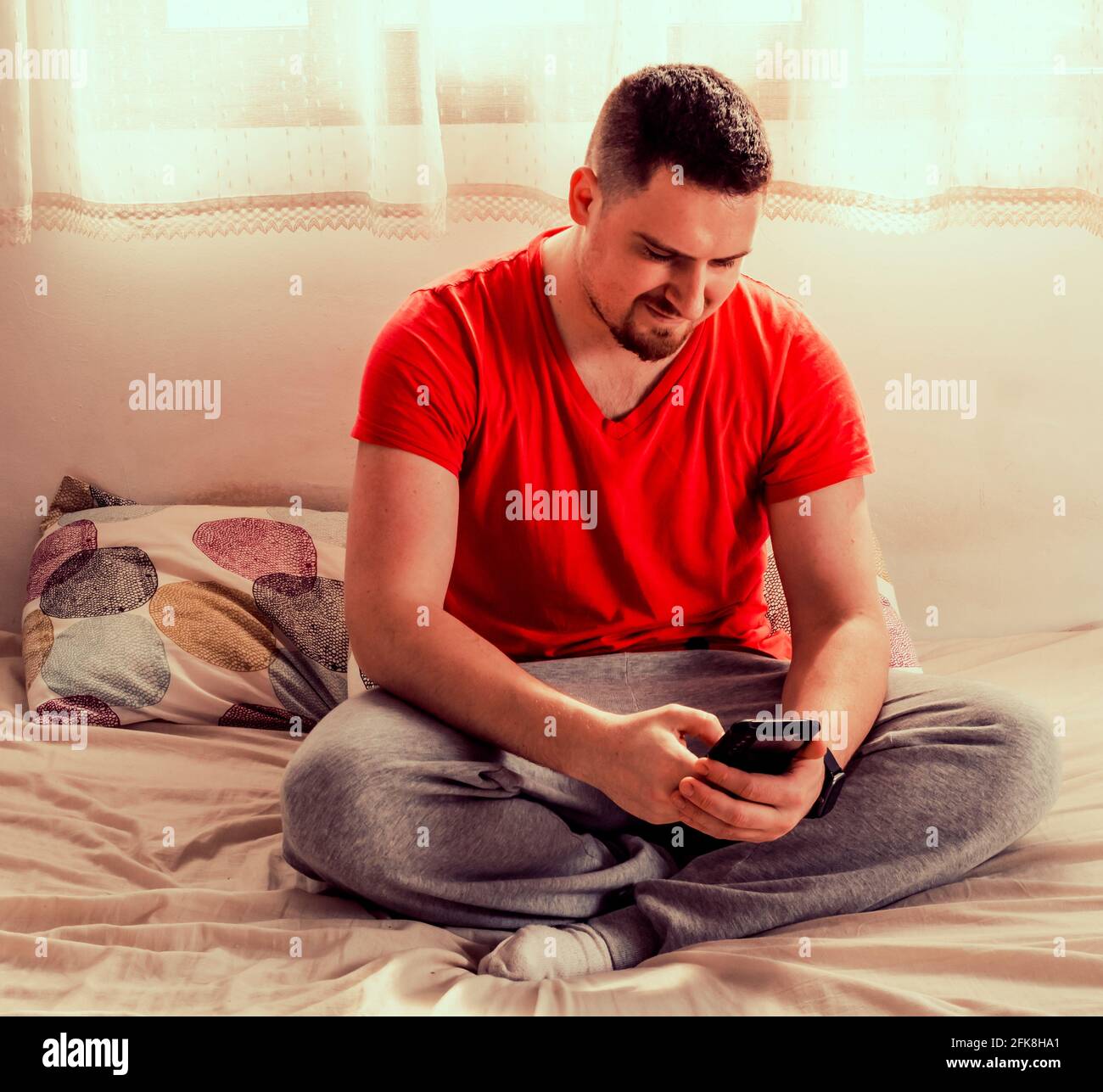 Bell'uomo sdraiato sul letto nella sua stanza indossando abiti casual, utilizzando uno smartphone Foto Stock