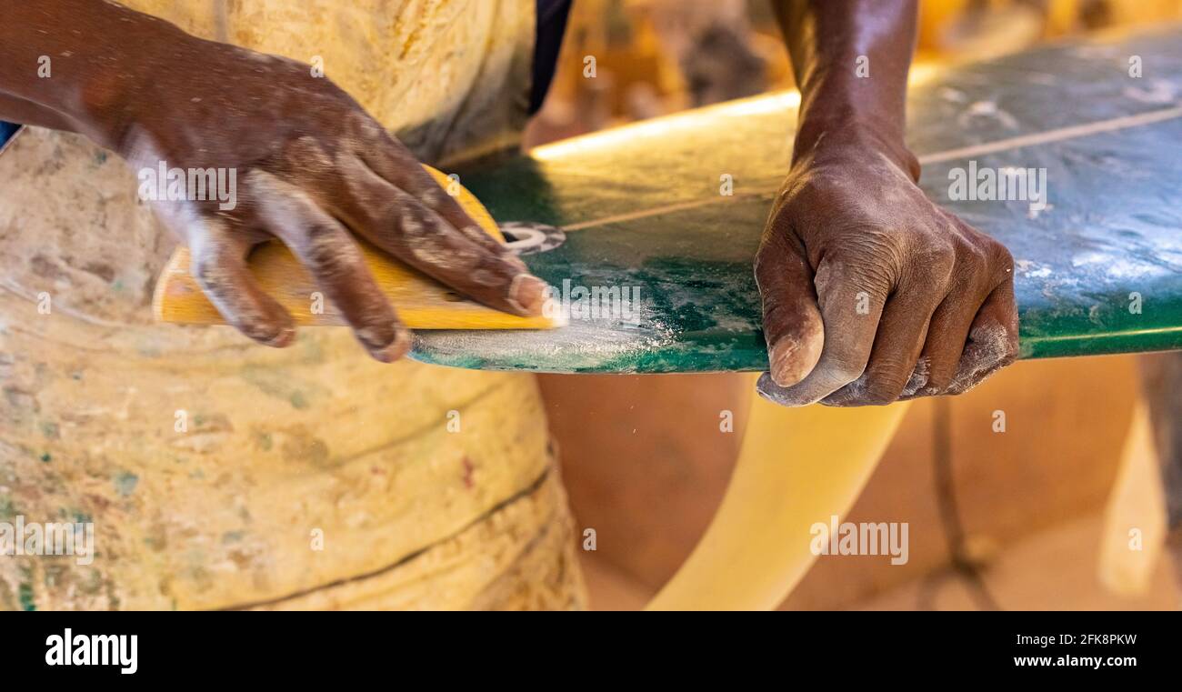 Uno Shaper da surf di un artigiano africano che lavora in un'officina di riparazione Con carta vetrata Foto Stock