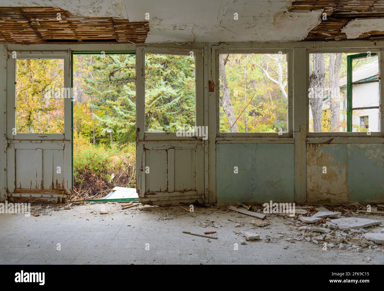 Casa abbandonata interno. Galleria con serramenti, vetri rotti e intonaco caduto, giardino d'autunno sopravorto visto attraverso le finestre Foto Stock