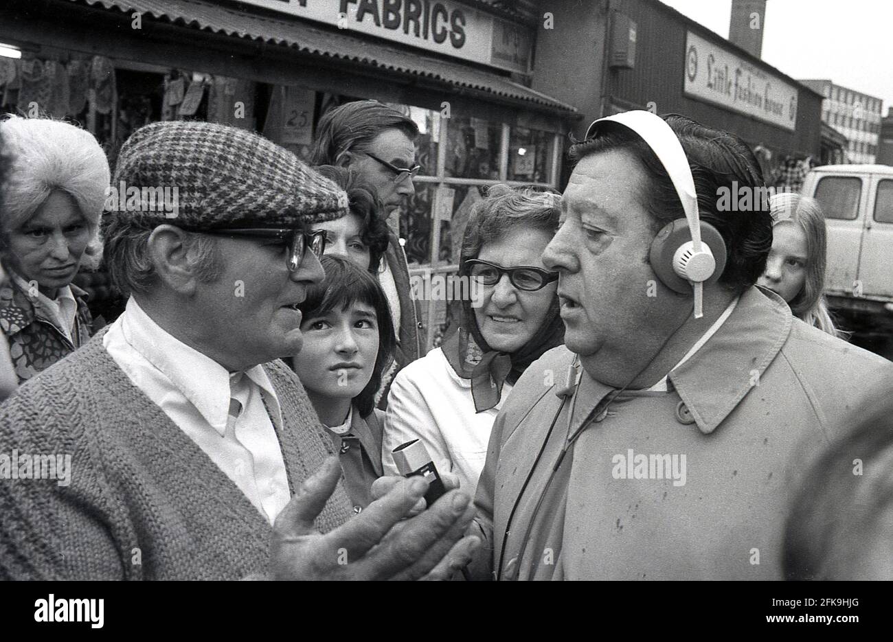 1970, uomo popolare della gente, presentatore radiofonico e reporter, Monty Modlyn parla con un uomo locale che indossa un cappello piatto circondato da persone in una zona di mercato del sud di Londra, Inghilterra, Regno Unito. Nato da una famiglia ebraica di Lambeth, Modlyn era noto per il suo accento di cetney e la sua trasmissione forte stava intervistando la gente britannica comune, una tecnica che divenne conosciuta come 'Vox Pop'. Rivolgendosi alle persone con le parole 'Ullo darlin', il suo personaggio della classe lavoratrice dell'estremità orientale lo ha visto parlare di tutto e di tutto durante una lunga e distinta carriera di radiodiffusione. Foto Stock