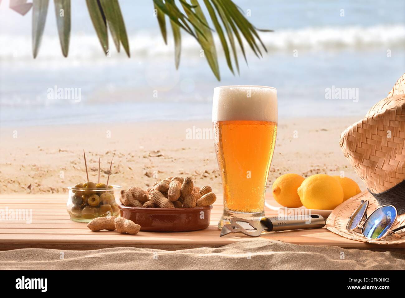 Dettaglio di un bicchiere di birra fresca su legno nel sabbia con un succulento spuntino su una spiaggia con albero di palma in una giornata calda Foto Stock