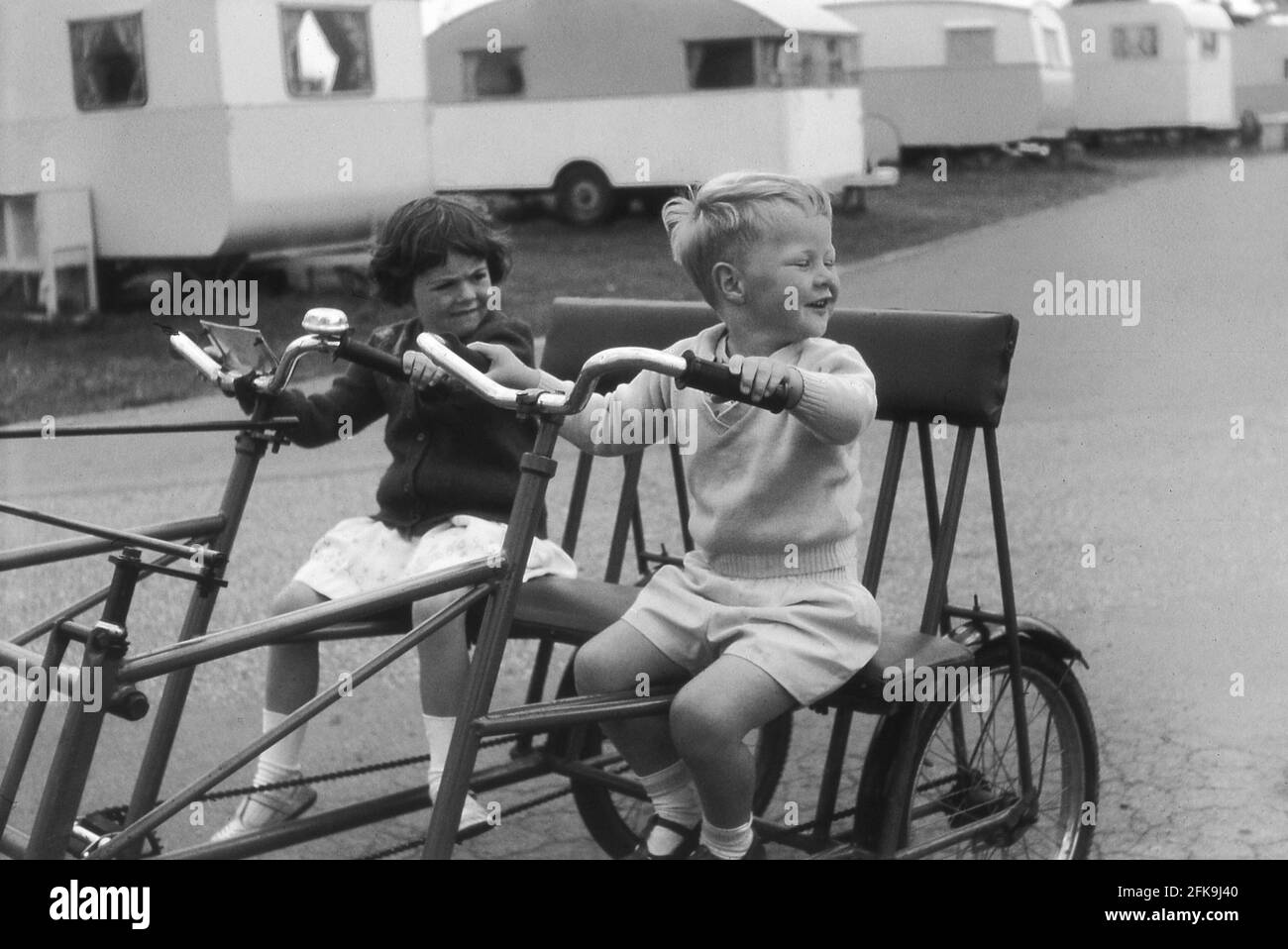1964, storico, fuori su un sentiero in un parco caravan vacanza, un ragazzo con la sua sorellina, seduto su una grande metallo incorniciato 'campo di vacanze' in bicicletta gemella o due posti, Suffolk, Inghilterra, Regno Unito. Queste biciclette a due posti sono state molto divertenti e un buon modo di muoversi intorno al sito sugli ampi sentieri. Foto Stock