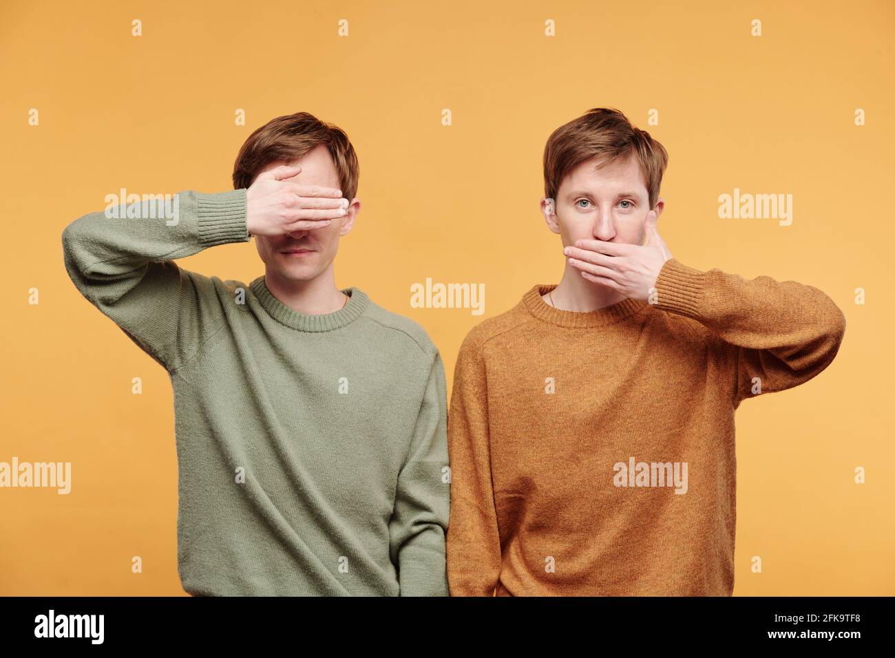 Ritratto di fratelli gemelli in maglioni in piedi su sfondo arancione e raffigurante scimmie saggi con occhi e bocca di copertura Foto Stock
