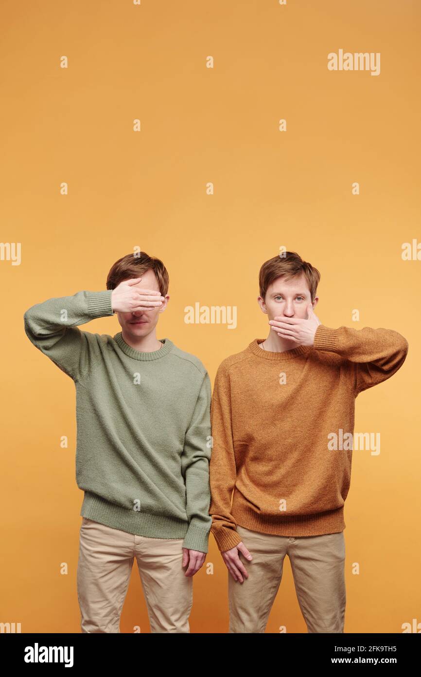 Un fratello gemello in maglione che copre gli occhi mentre l'altro fratello bocca di copertura su sfondo arancione Foto Stock