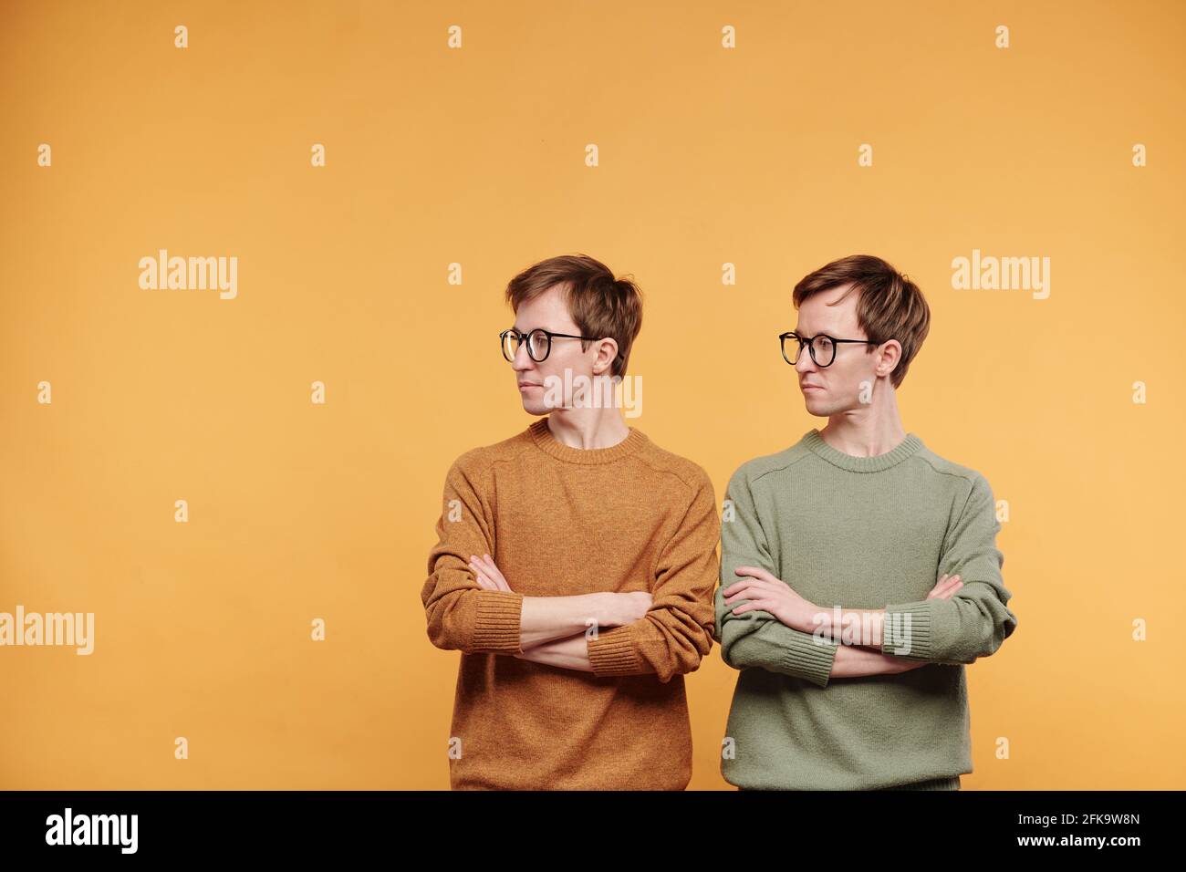 Gemelli rigidi in occhiali e pullover luminosi in piedi con braccia incrociate e guardando da parte, sfondo arancione Foto Stock