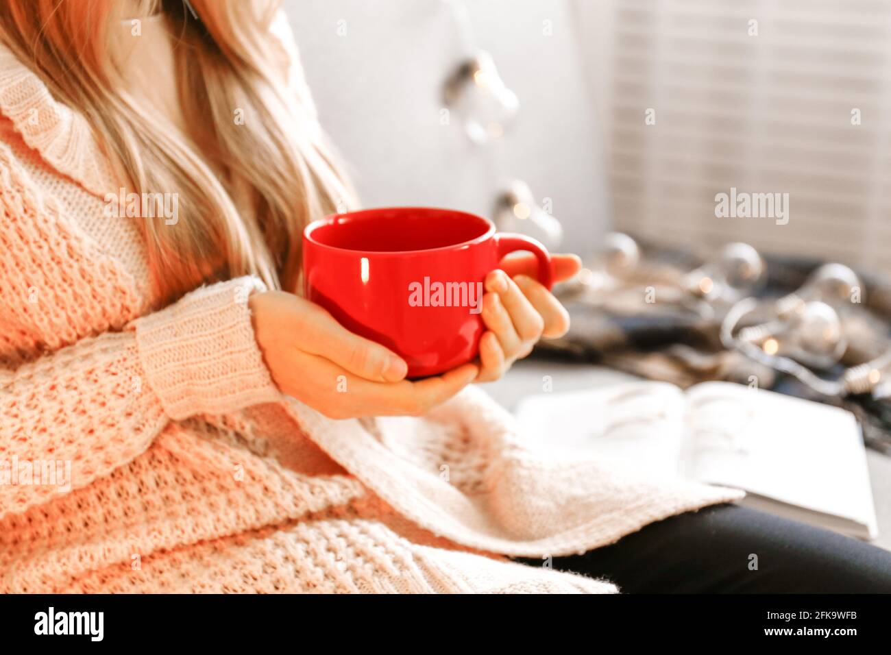 Primo piano le mani di una giovane donna che tiene una grande tazza di bevanda calda. Donna che indossa un pullover in maglia bianca oversize alla moda, seduto a casa con tazza di cof Foto Stock