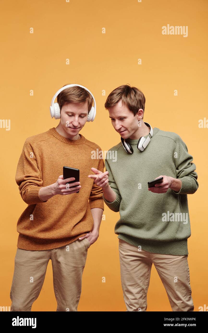 Giovane uomo rosso in cuffie wireless che mostrano lo schermo del telefono e. condivisione di brani musicali con il fratello gemello Foto Stock