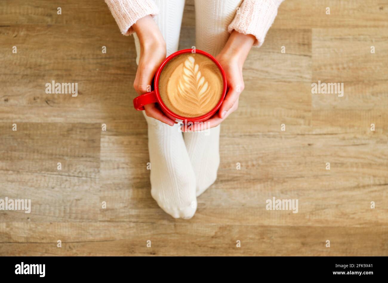 Giovane donna che beve il cappuccino e si siede sul pavimento di legno. Vista dall'alto delle gambe femminili in calze calde bianche. Vacanze invernali comfort Foto Stock