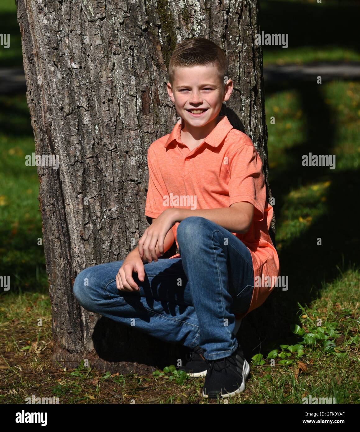 Il ragazzo dà un grin boyish mentre inginocchiandosi oltre ad un albero nel sole di mattina. Indossa jeans e una camicia in stile polo arancione. Foto Stock