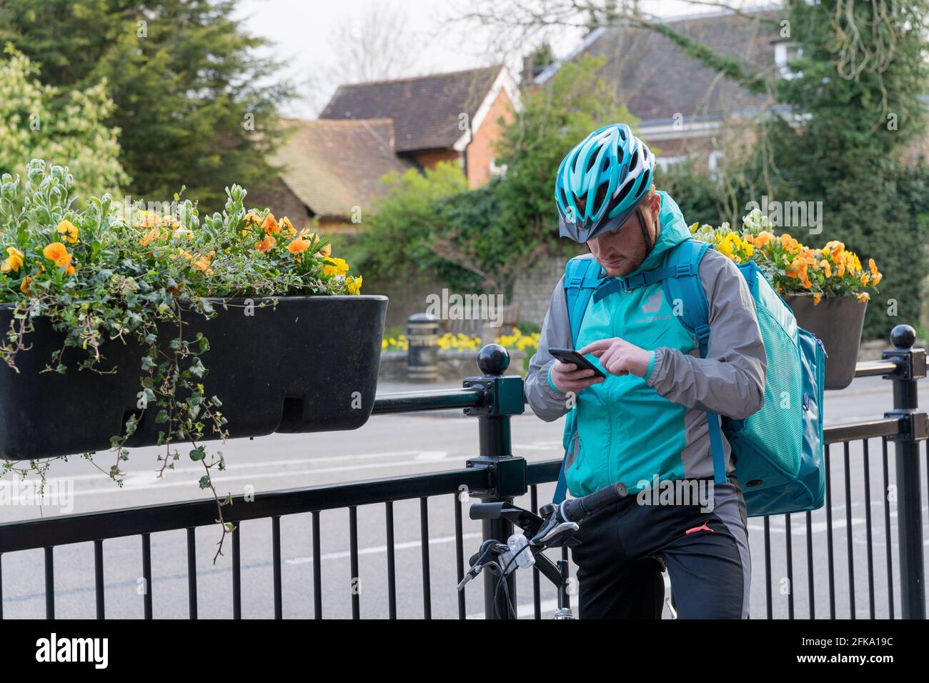 Il biker di Deliveroo controlla il suo mobile per la direzione all'indirizzo del cliente sulla plavement dalle ringhiere di ferro con cesto di fiori appesi, Kent Foto Stock