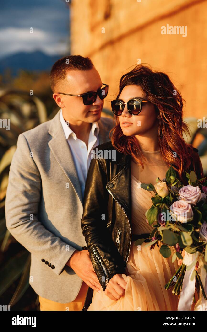 Lo sposo si trova dietro la sposa in occhiali da sole e una giacca in pelle  con un bouquet di fiori sullo sfondo del muro del castello Foto stock -  Alamy