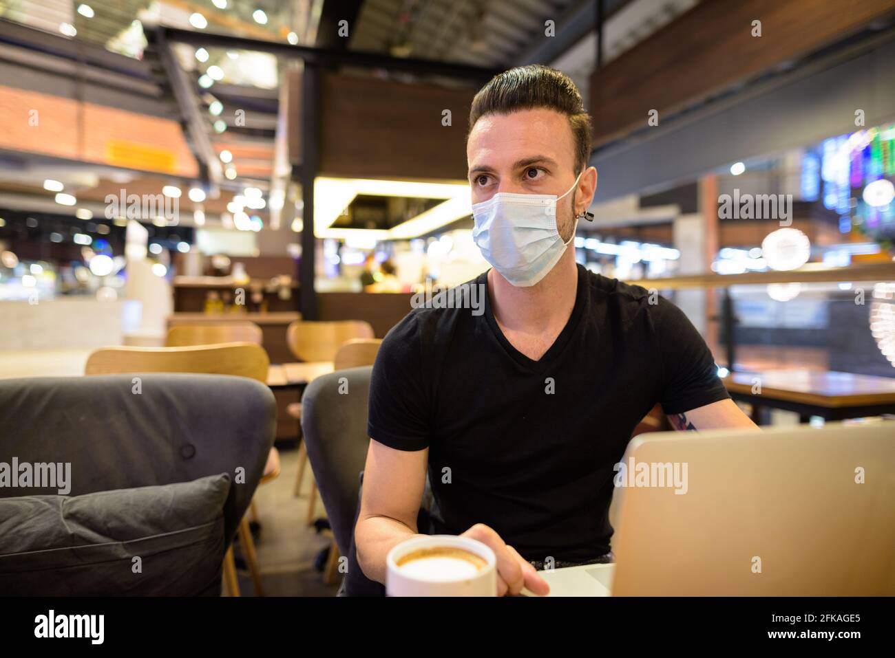 Uomo serio seduto al bar che indossa la maschera facciale e. utilizzare un computer portatile mentre si pensa Foto Stock