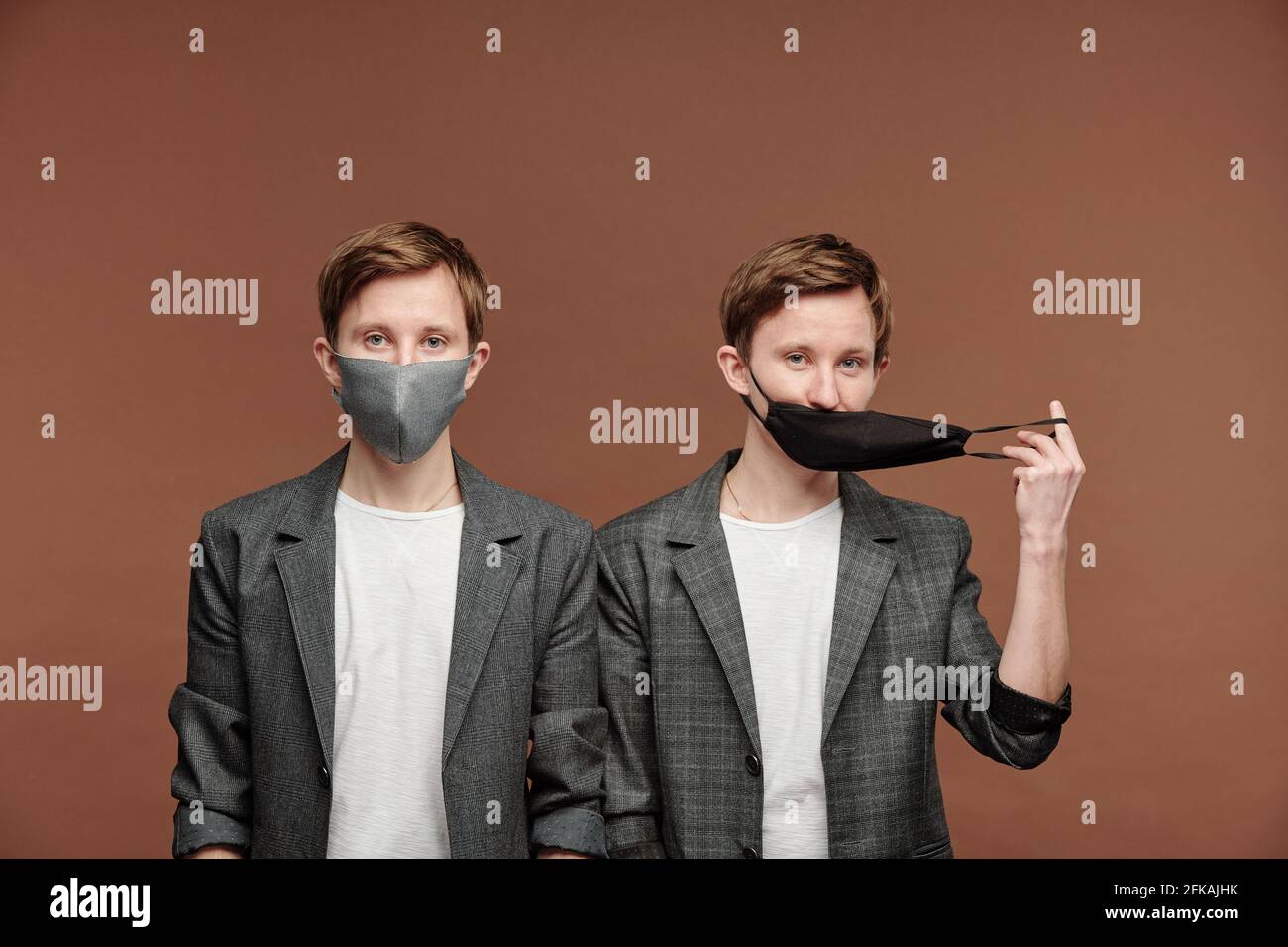 Ritratto di bei fratelli gemelli in eleganti abiti grigi che si posano in maschere su sfondo marrone, uno di loro tirando maschera Foto Stock