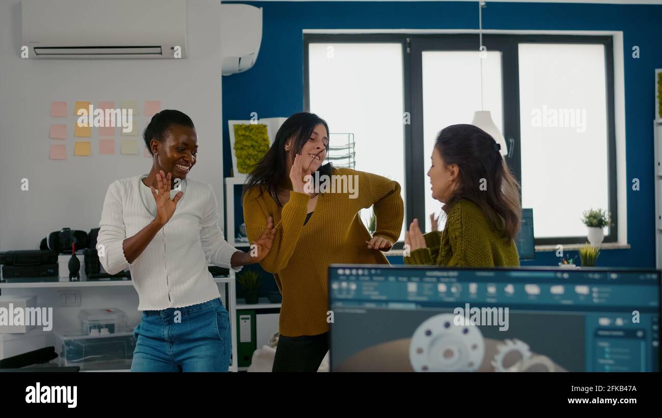 Entusiaste donne di ingegneri diversi che ballano in ufficio aziendale celebrando il successo del progetto, cantando e divertendosi durante l'orario di lavoro. Ha entusiasmato i progettisti multietnici che utilizzano un pc che mostra il software cad Foto Stock