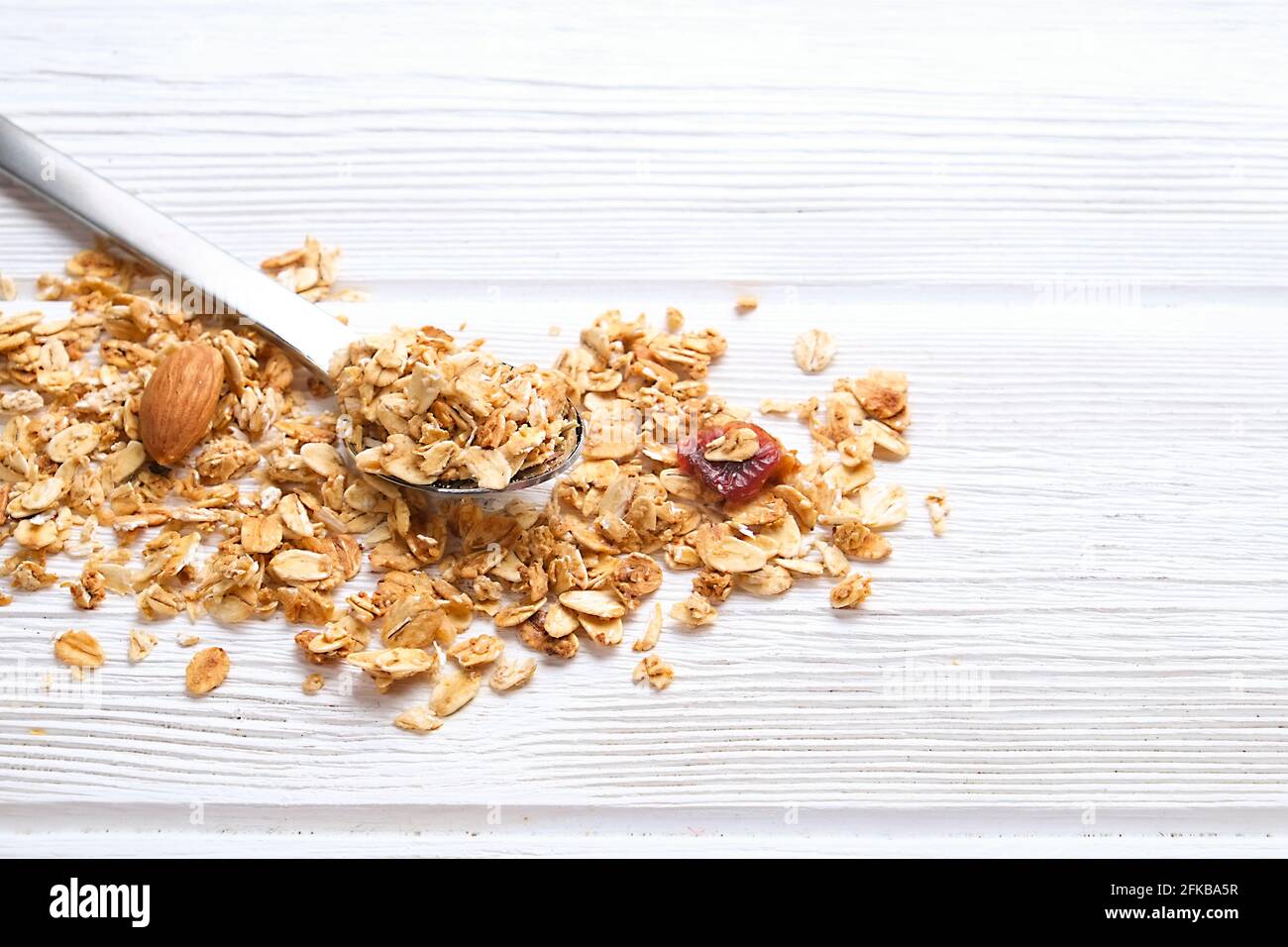 Cucchiaio di avena arrotolata sparsa di granola su tavola di tessitura in legno bianco. Fiocchi vegani nutrienti sani pieni di carboidrati e proteine per s. Foto Stock