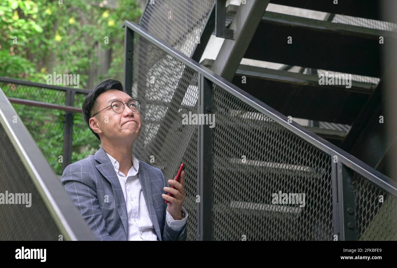 Uomo d'affari asiatico in costume con gli occhi guardando in su, pensando a qualcosa. Foto Stock