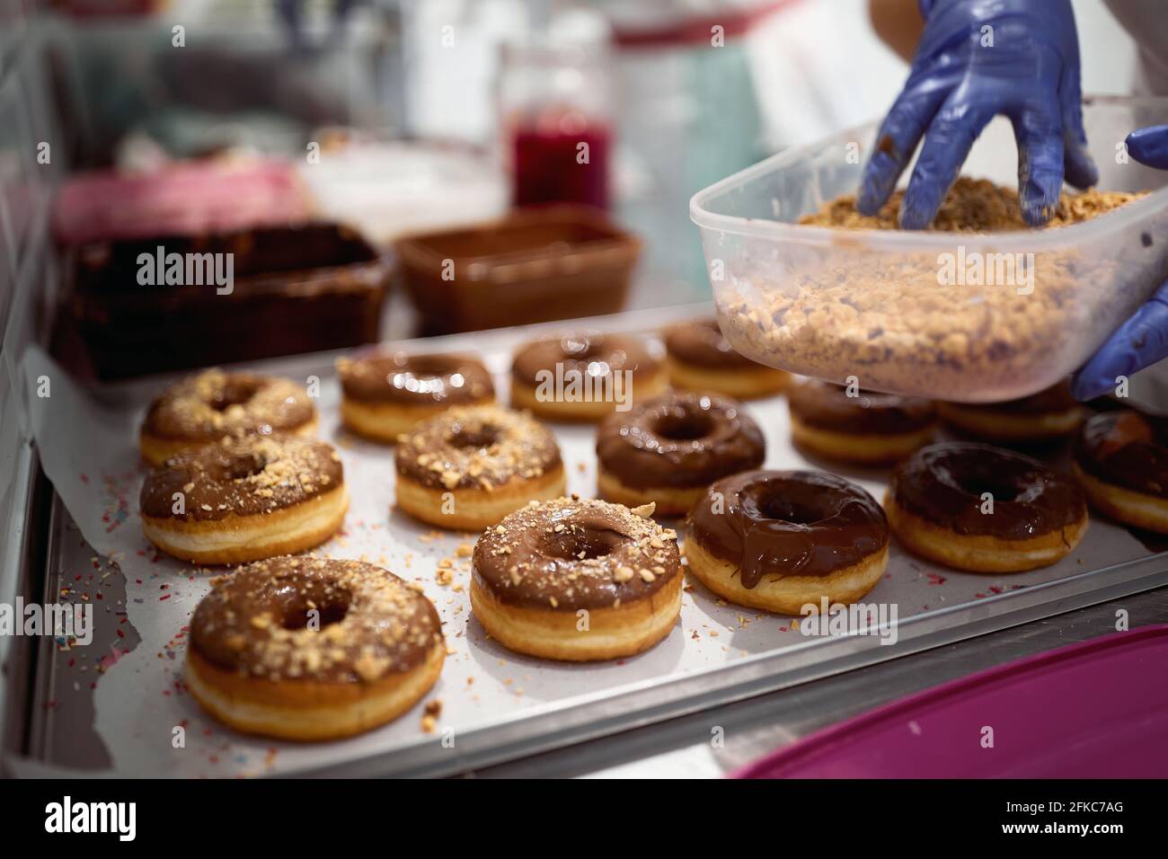 Un lavoratore spruzza i biscotti schiacciati sopra le ciambelle deliziose in un'atmosfera di lavoro in un laboratorio di caramelle. Pasticceria, dessert, dolce Foto Stock