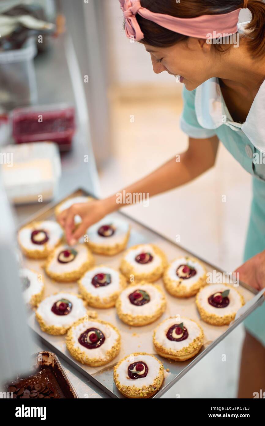 Un giovane proprietario femminile di piccola impresa sta decorando le sue deliziose ciambelle fatte a mano di aspetto irresistibile. Pasticceria, dessert, dolce, fare Foto Stock