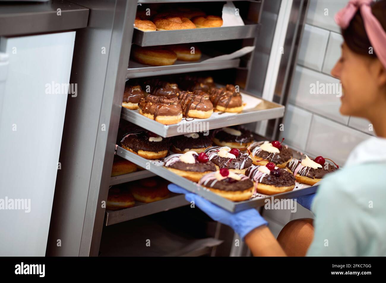 Una giovane piccola donna di affari riempie l'armadio con deliziose ciambelle fatte a mano in un'atmosfera piacevole in un laboratorio di caramelle. Pasticceria, dessert, swe Foto Stock