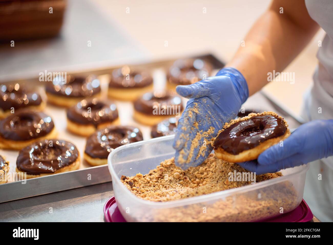 Un lavoratore spruzza nocciole macinate su ciambelle in un'atmosfera di lavoro in un laboratorio di caramelle. Pasticceria, dessert, dolce, fare Foto Stock
