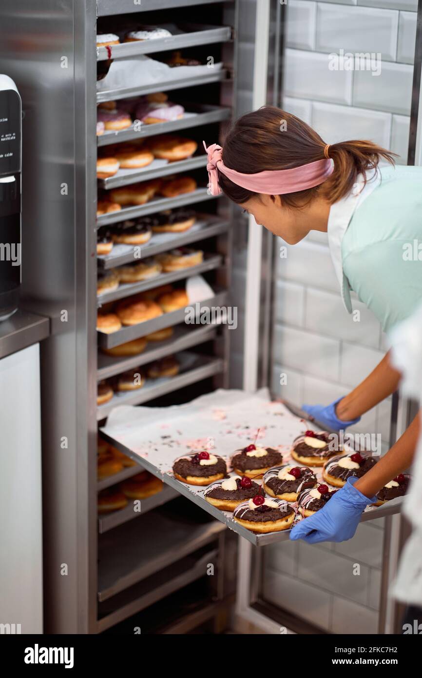 Una giovane piccola impresa donna proprietario sta imballando le ciambelle squisite fatte a mano pronte ad un vassoio in un'atmosfera di lavoro in un laboratorio di caramelle. Pasticceria, dessert, Foto Stock