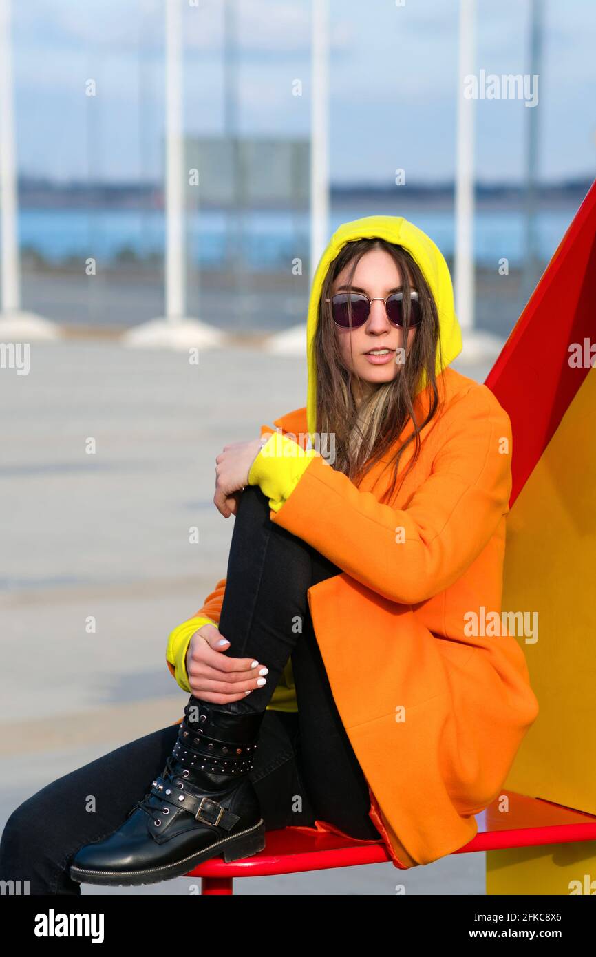 Millennial Hipster Girl in occhiali da sole lilla, cappotto arancione,  felpa con cappuccio gialla in posa