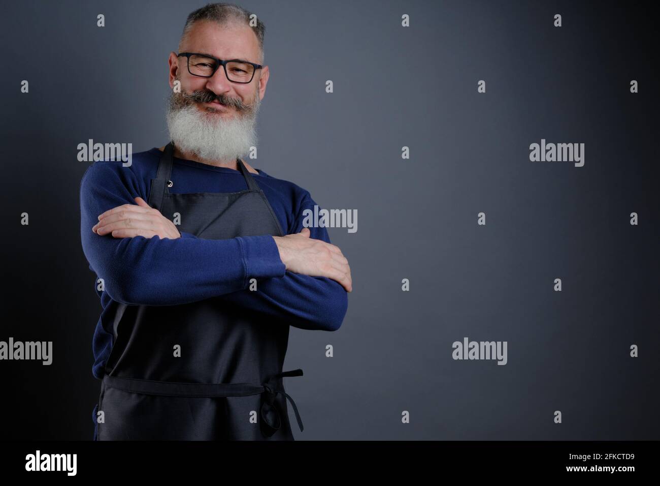 Ritratto di un bel grembiule maturo isolato su sfondo grigio, uomo caucasico con barba sorridente Foto Stock