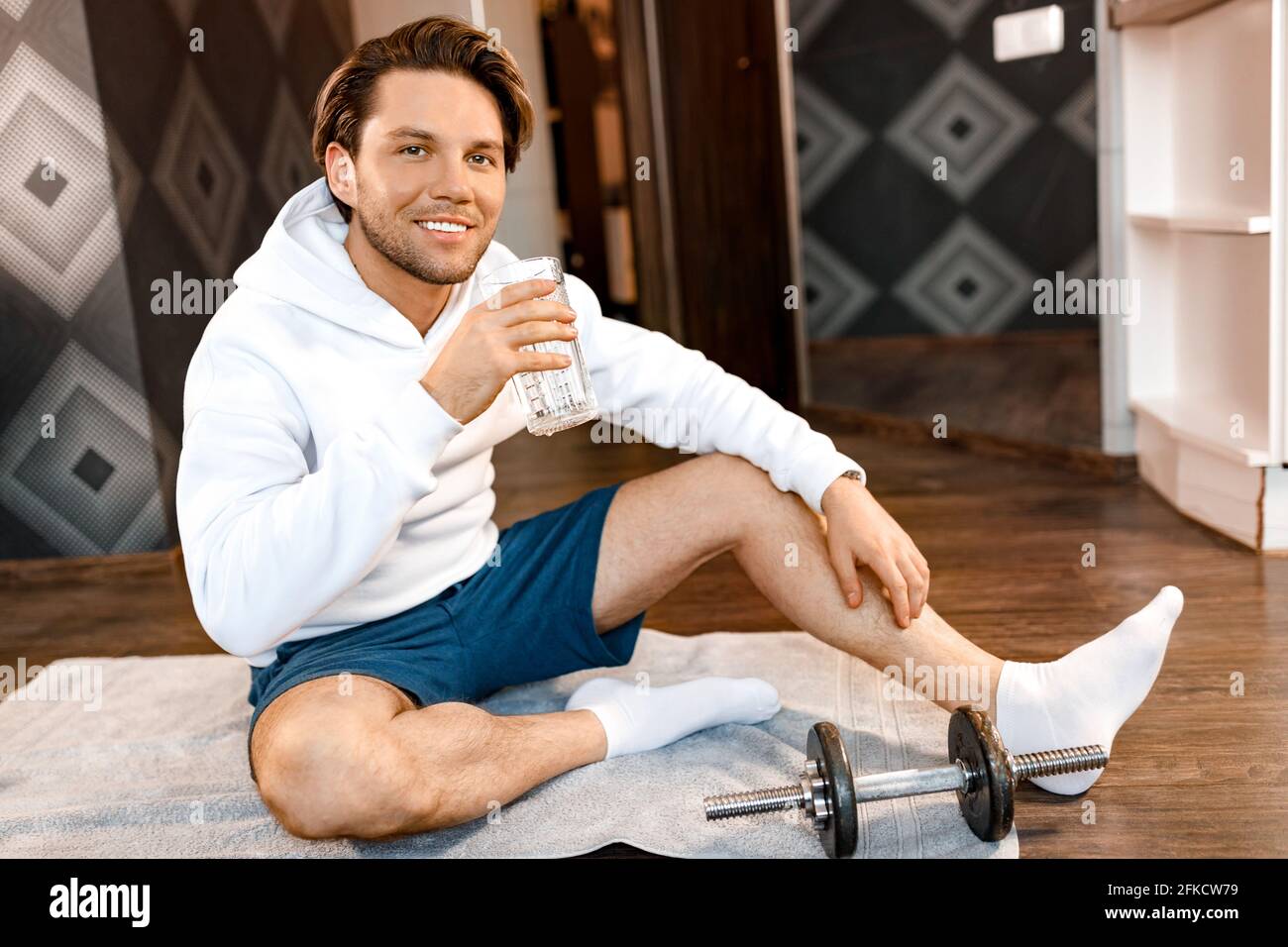 Bell'uomo elegante seduto sul tappetino dopo l'allenamento, guardando la fotocamera, sorridendo. Il maschio atletico che tiene un bicchiere di acqua pulita, segue sano Foto Stock