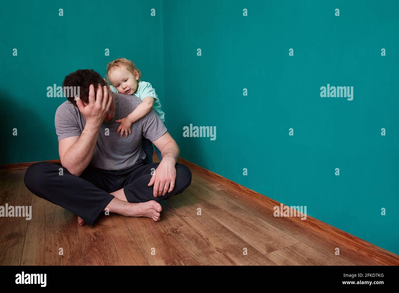 Il bambino si sente dispiaciuto per il padre sconvolto seduto sul pavimento in una camera verde cupa. Spazio di copia. Foto Stock